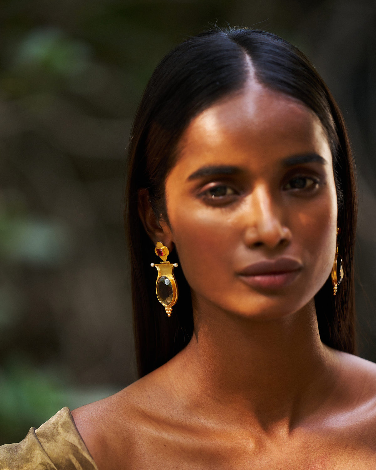 Person wearing gold eden drop earrings with red studs and oval glass.
