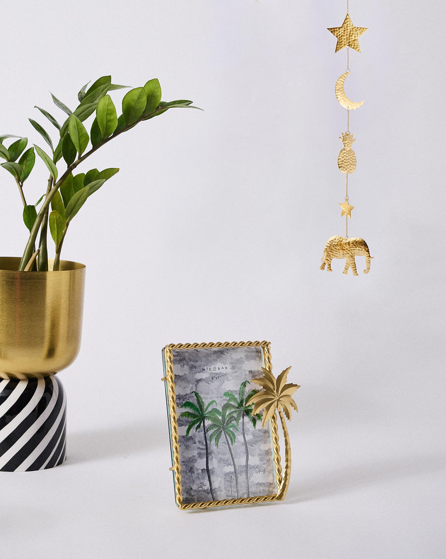 The Gilded Palm frame beside a plant in a gold pot and a hanging gold charm