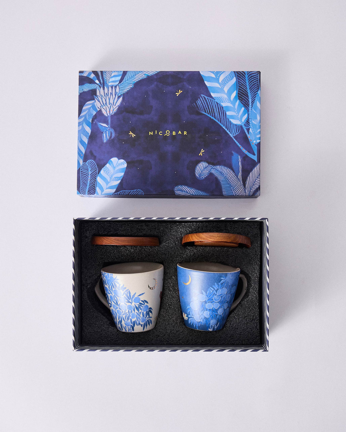 Open Nadi box with cream and blue botanical mugs, wooden lids, and leaf-print lid.