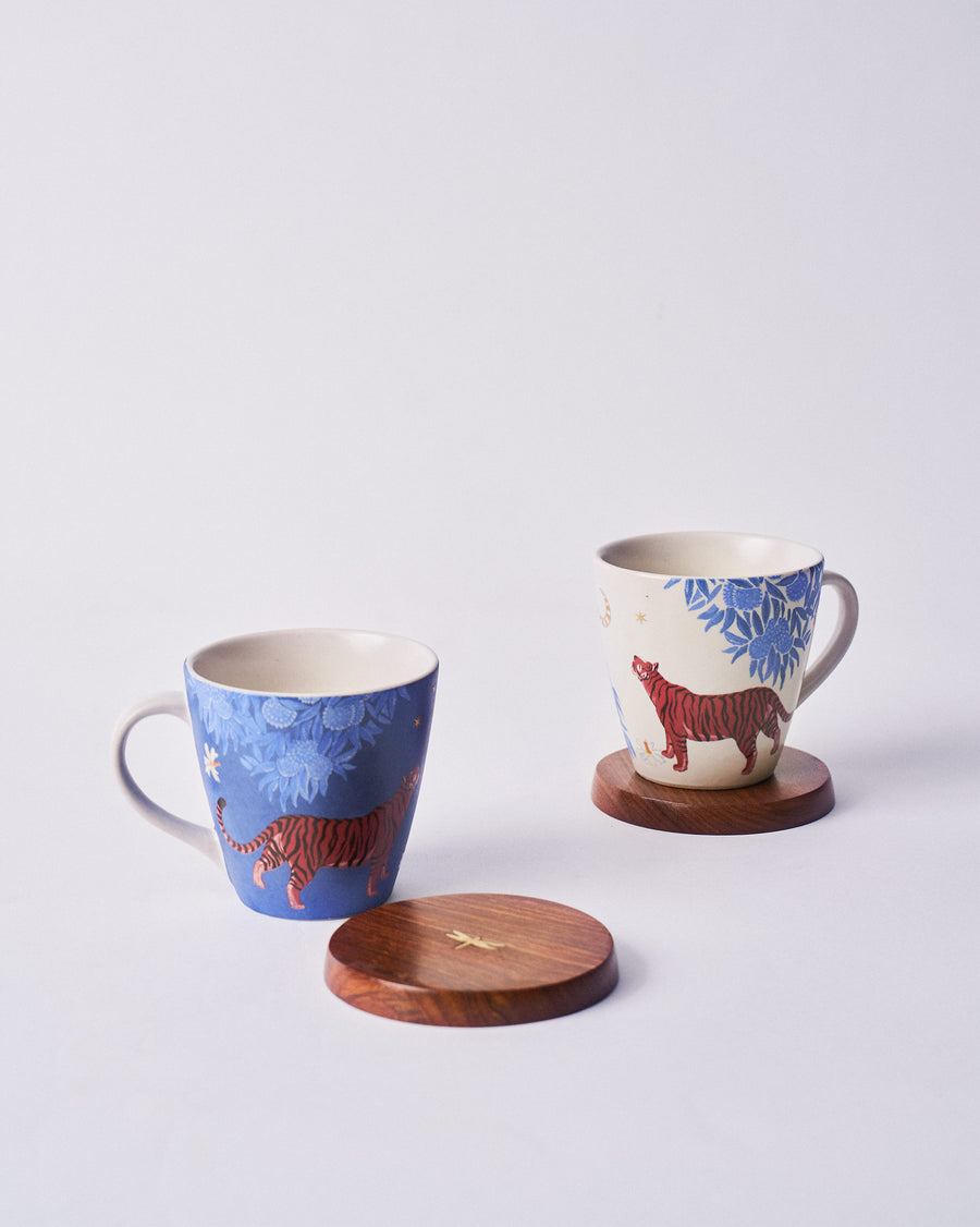 A blue Nadi mug and a cream one, each with a red tiger, beside a wooden lid
