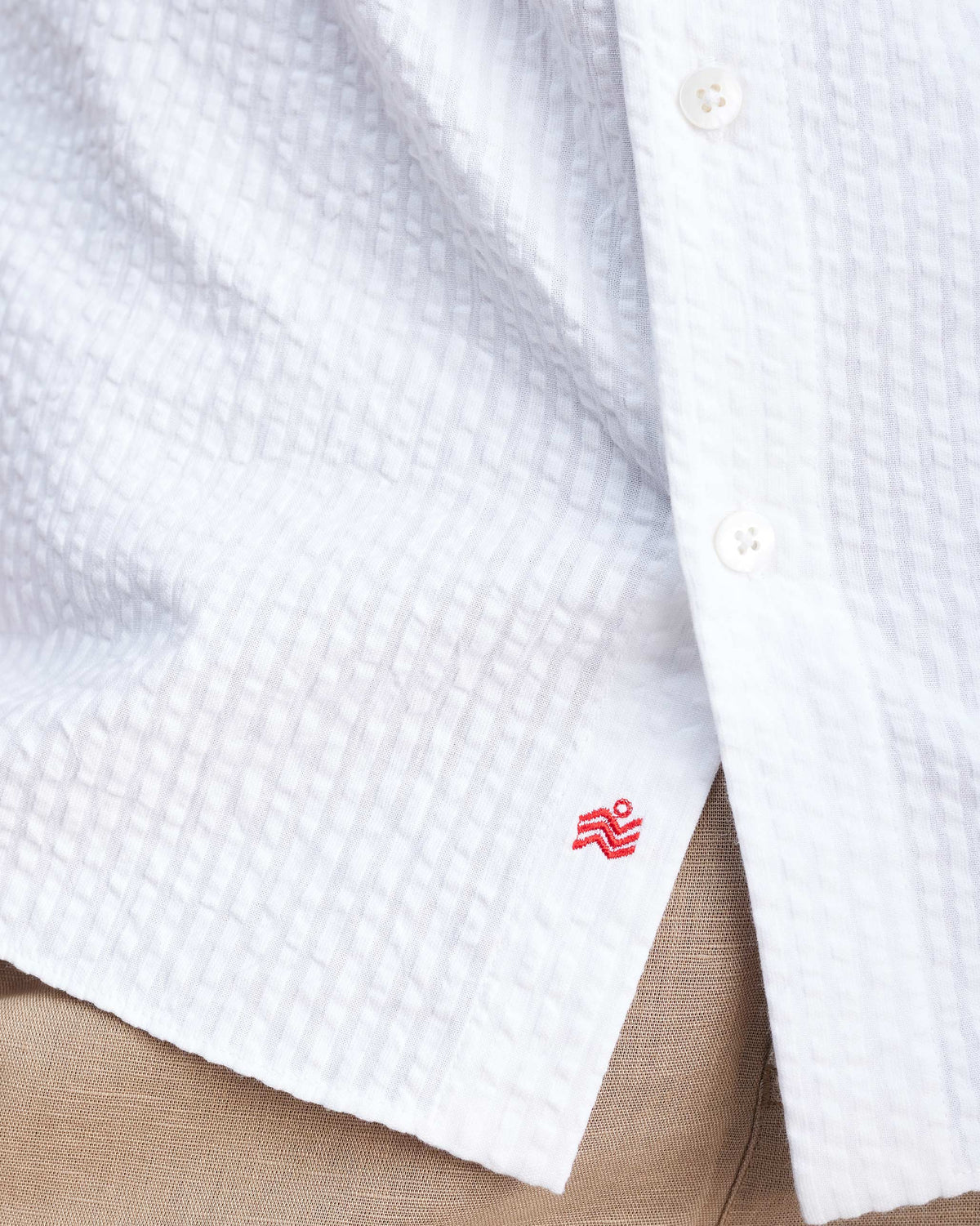 Close-up of a red logo near the hem of a white seersucker shirt.