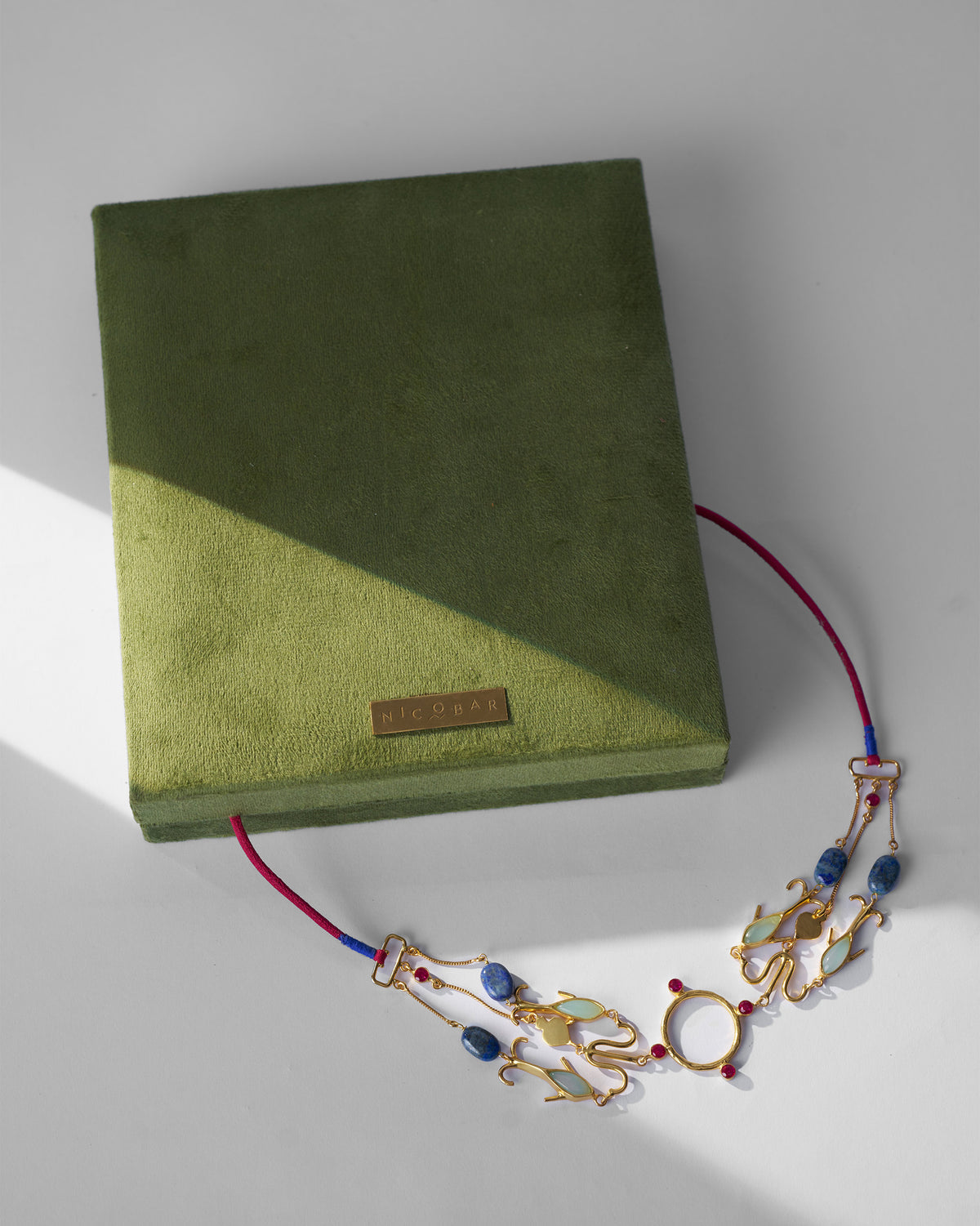 Gold Indus necklace with blue and green stones draped on an olive Nicobar box.