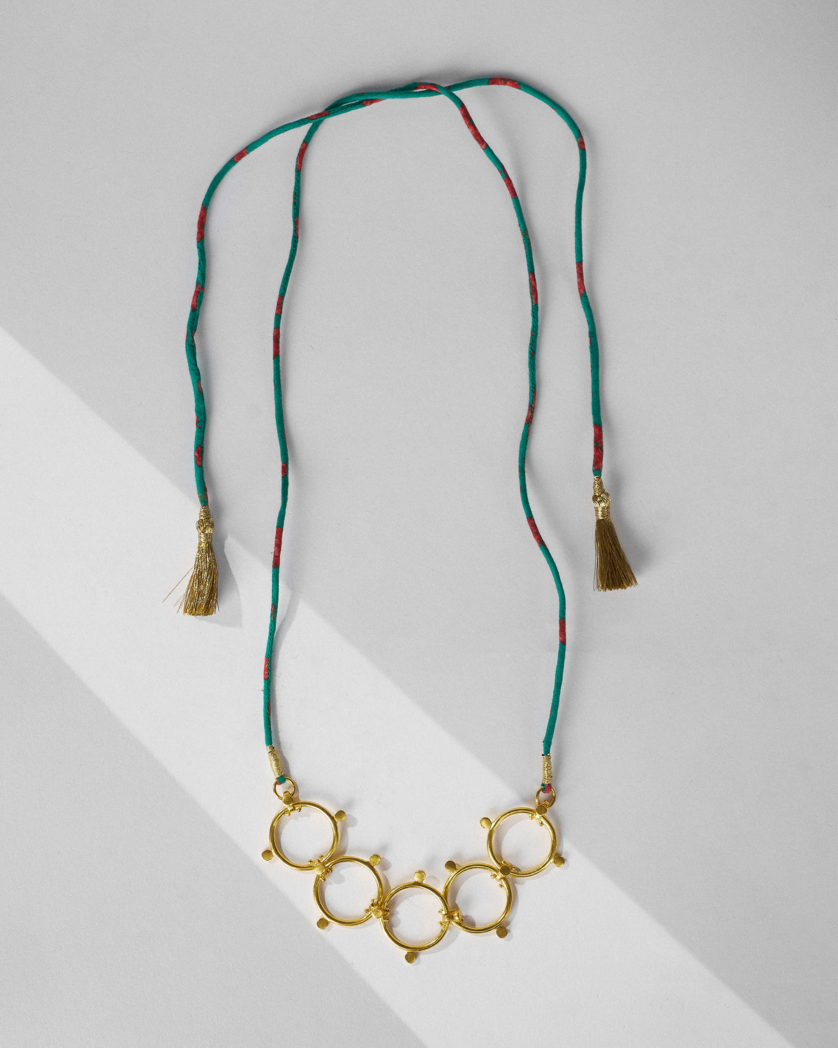 Full length of a Panch Circle gold necklace on a teal cord with tassels.