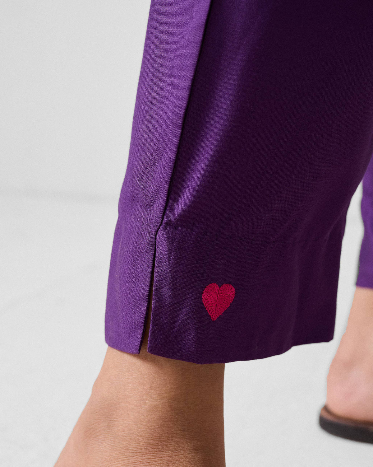 Close-up of a fuchsia heart embroidered near the hem of purple Darjeeling pants.
