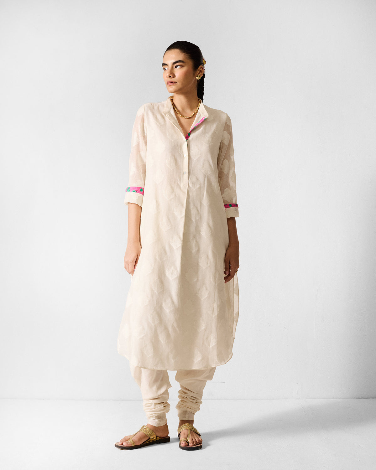 Person wearing an ivory long kurta with floral buta and pink-teal cuff trim.