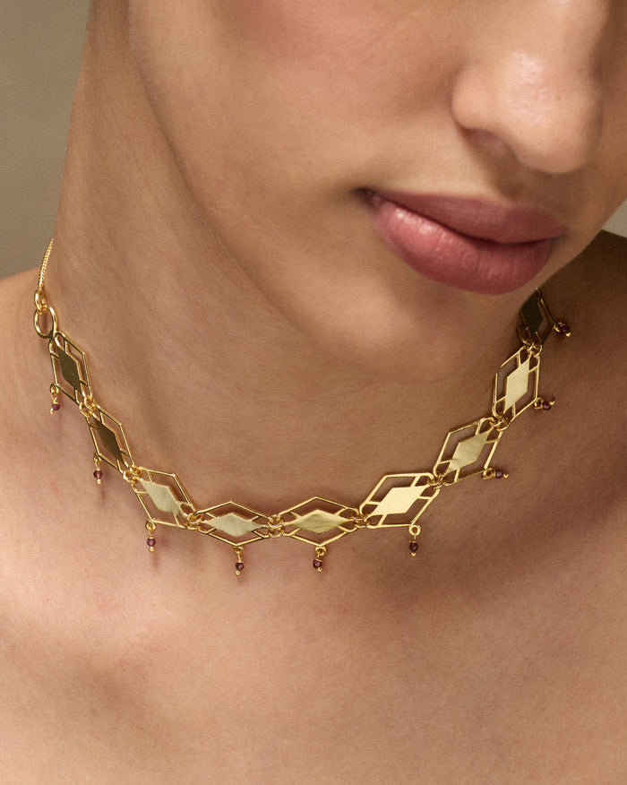 Gold diamond-link necklace with small red garnet drops worn at the neck.