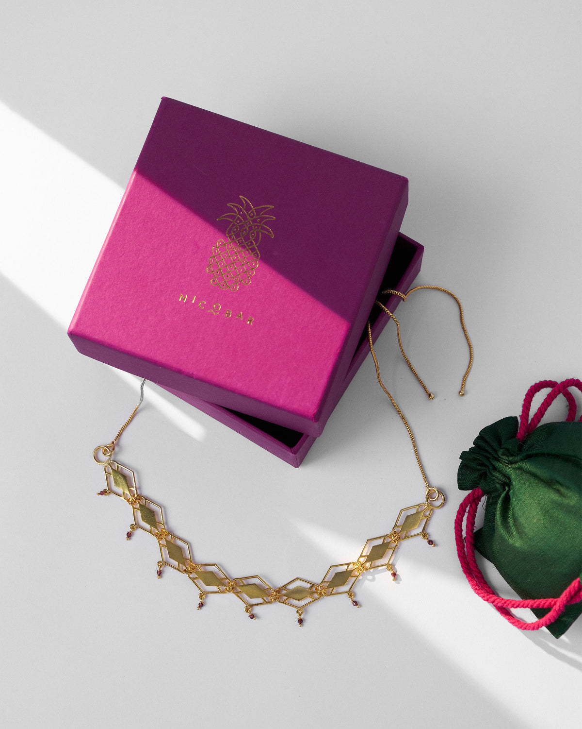 Gold barfi necklace with garnet drops beside a fuchsia Nicobar gift box.