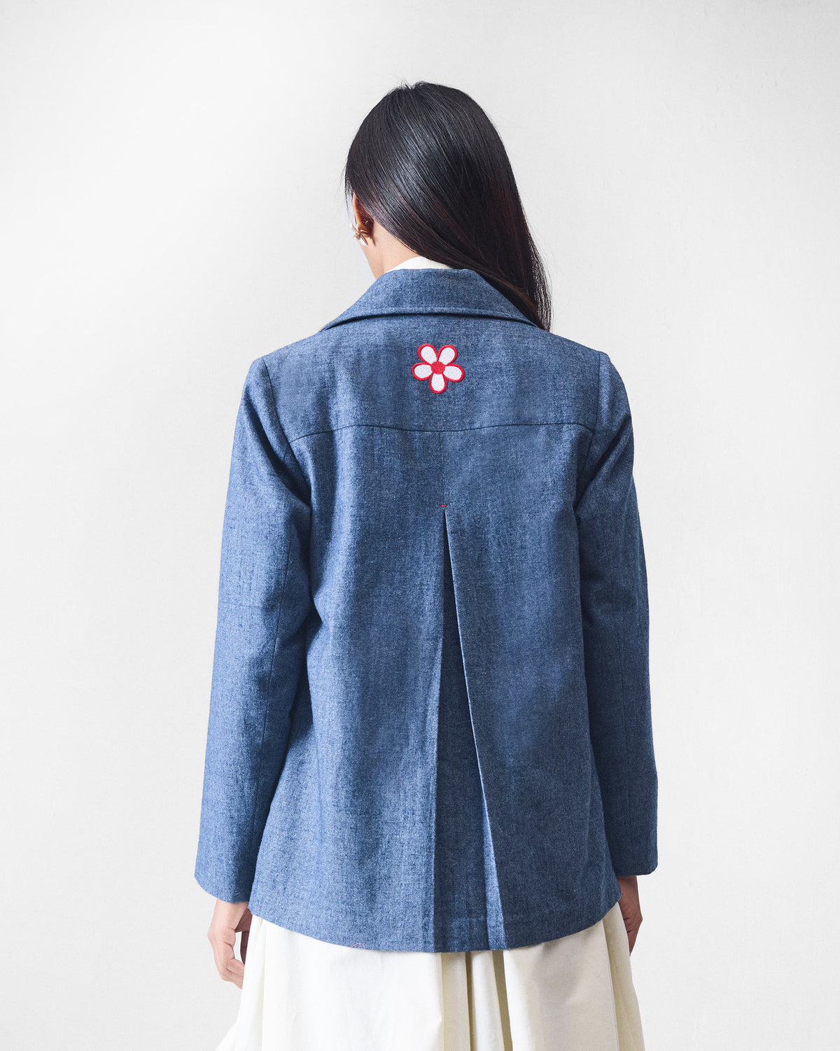 Back of a dust blue Trace Jacket with a center pleat and flower embroidery.
