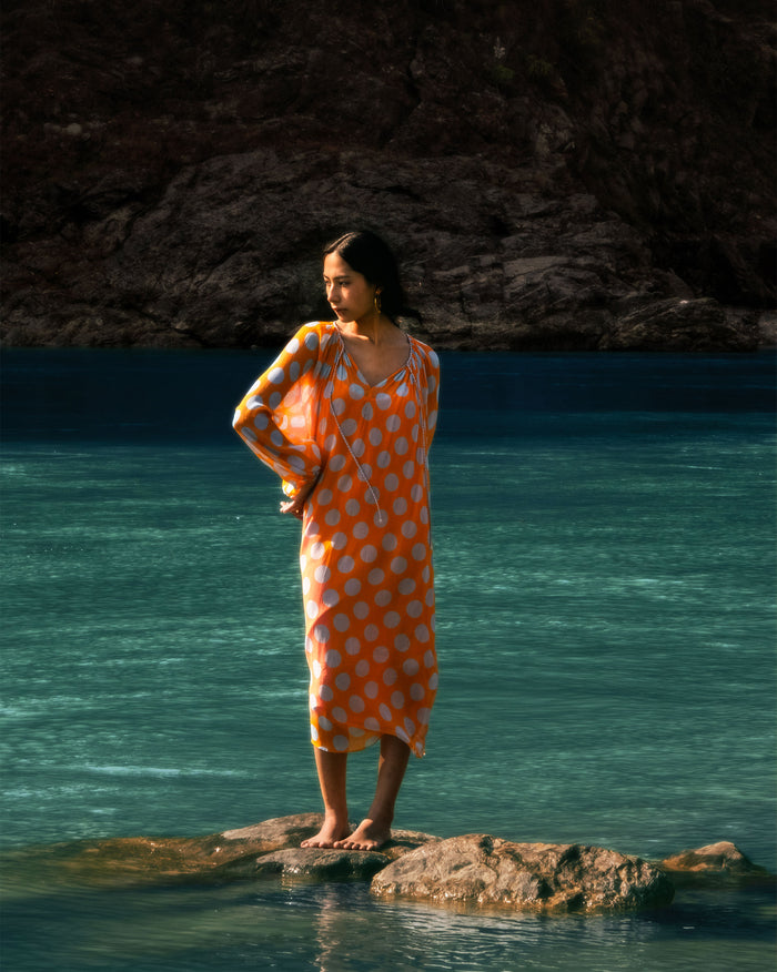 Person wearing an orange Sara kaftan with white dots by the water.