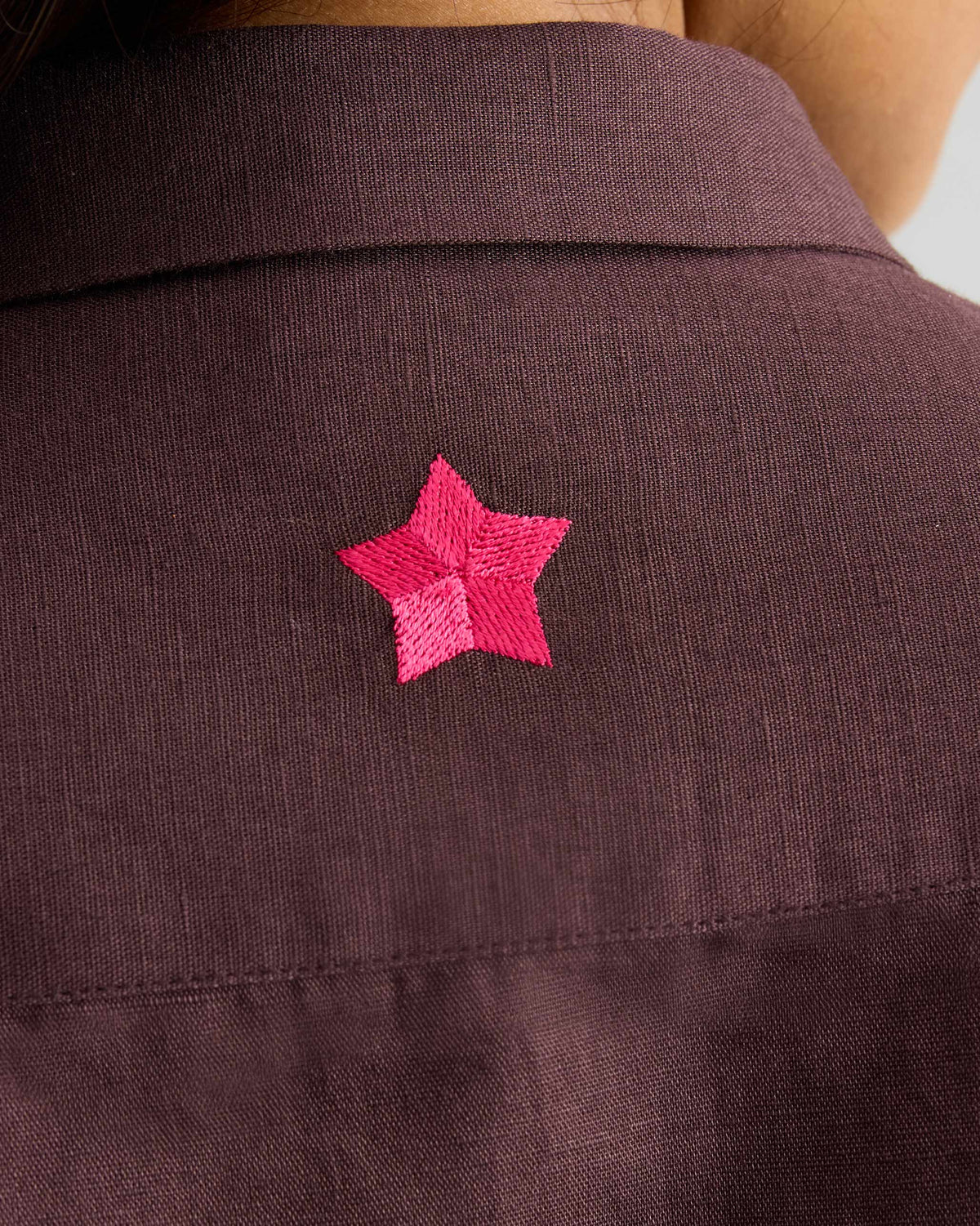 Close-up of a pink star embroidery on a chocolate plum Drop Armhole Shirt.