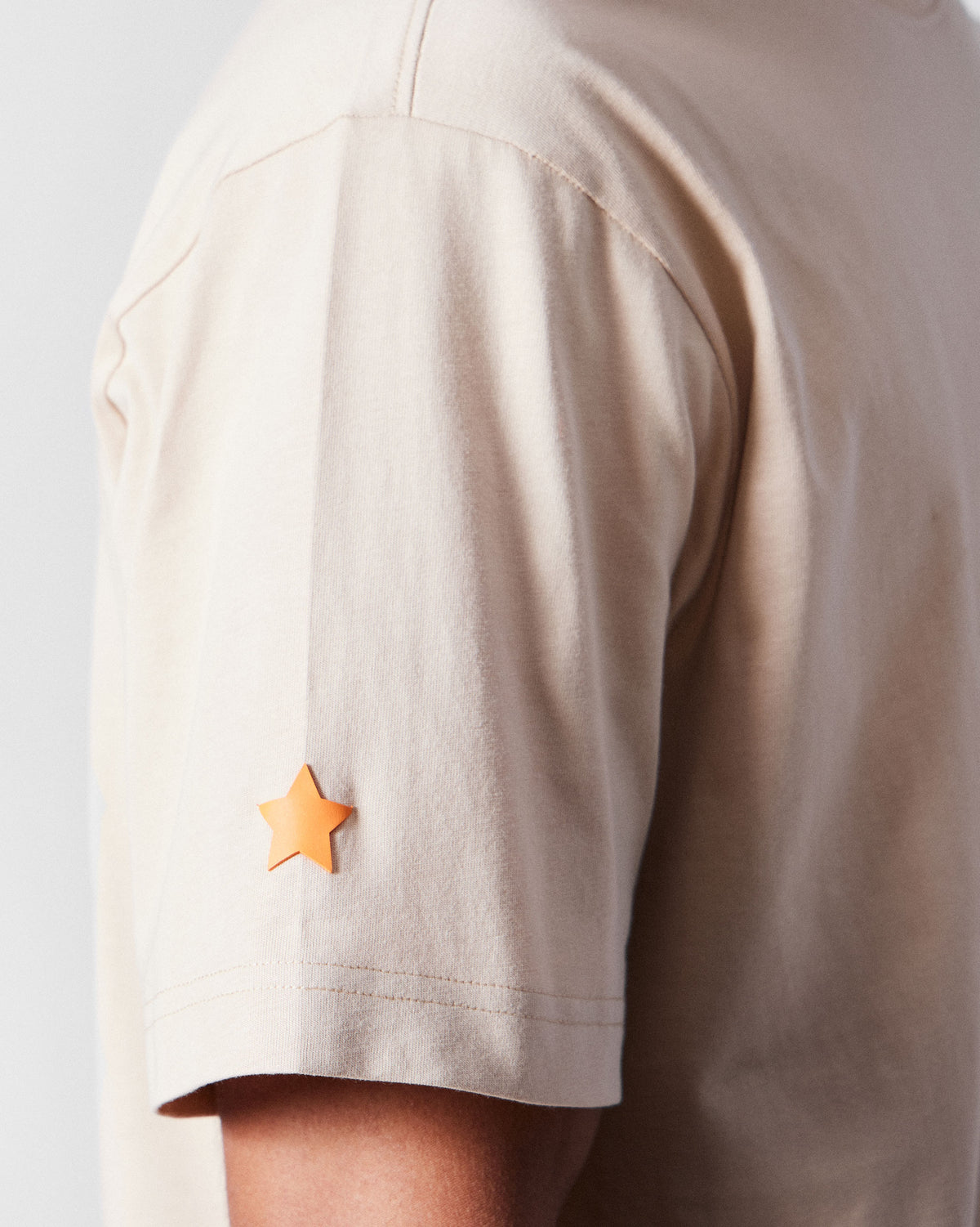 Close-up of an orange star on the sleeve of a beige Postal T-Shirt.
