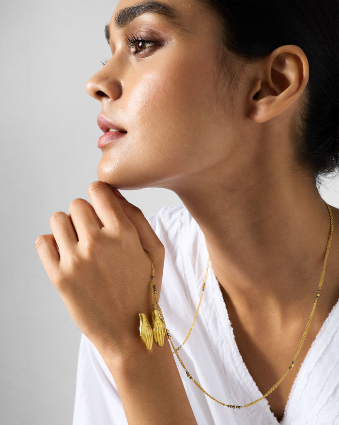 Gold Sama necklace with a prayer-hands pendant and dark beads.