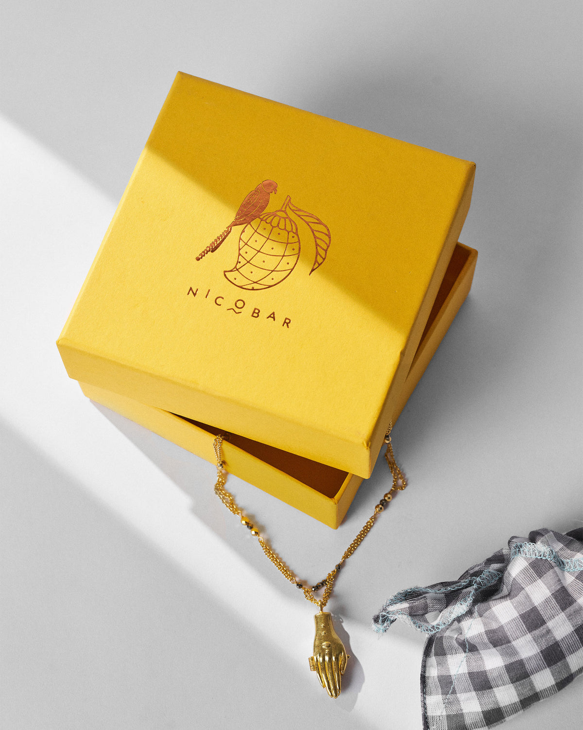 Gold Sama necklace with a hand pendant beside a yellow Nicobar gift box.