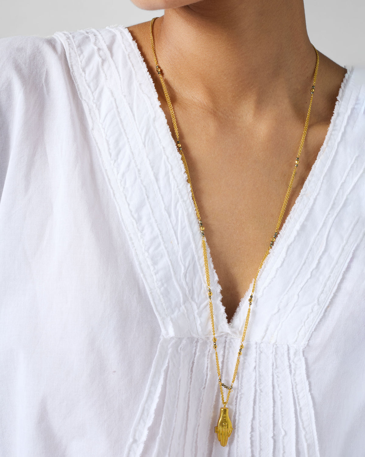 Long gold Sama necklace with a hand pendant worn over a white top.
