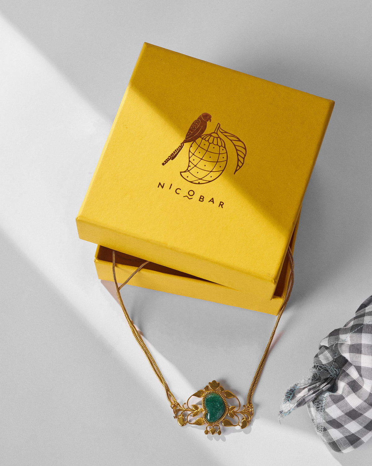 Gold Ambi choker with green stone displayed beside a yellow Nicobar gift box.
