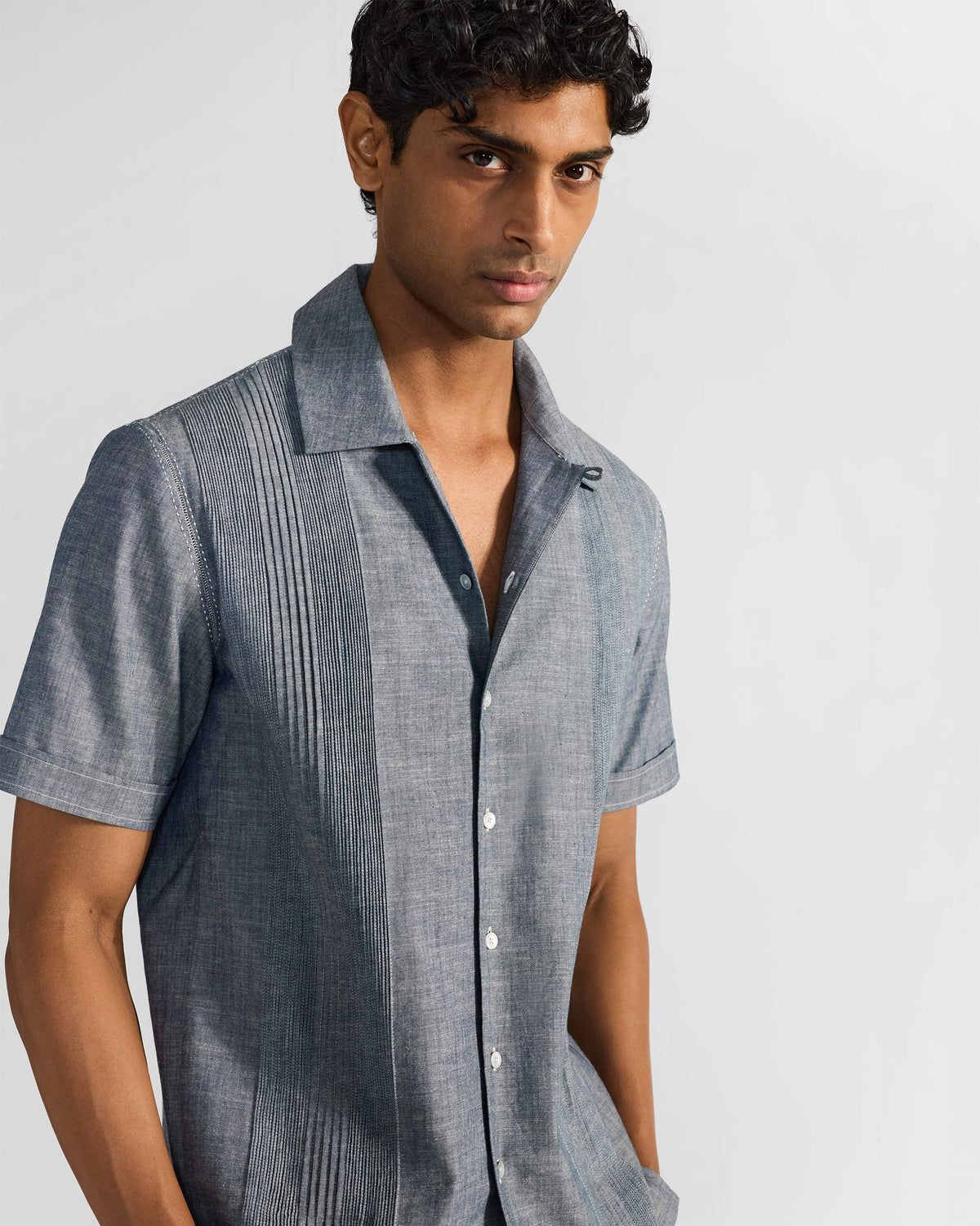 Man in an indigo short-sleeve shirt with front pintucks and a camp collar against a plain studio backdrop.