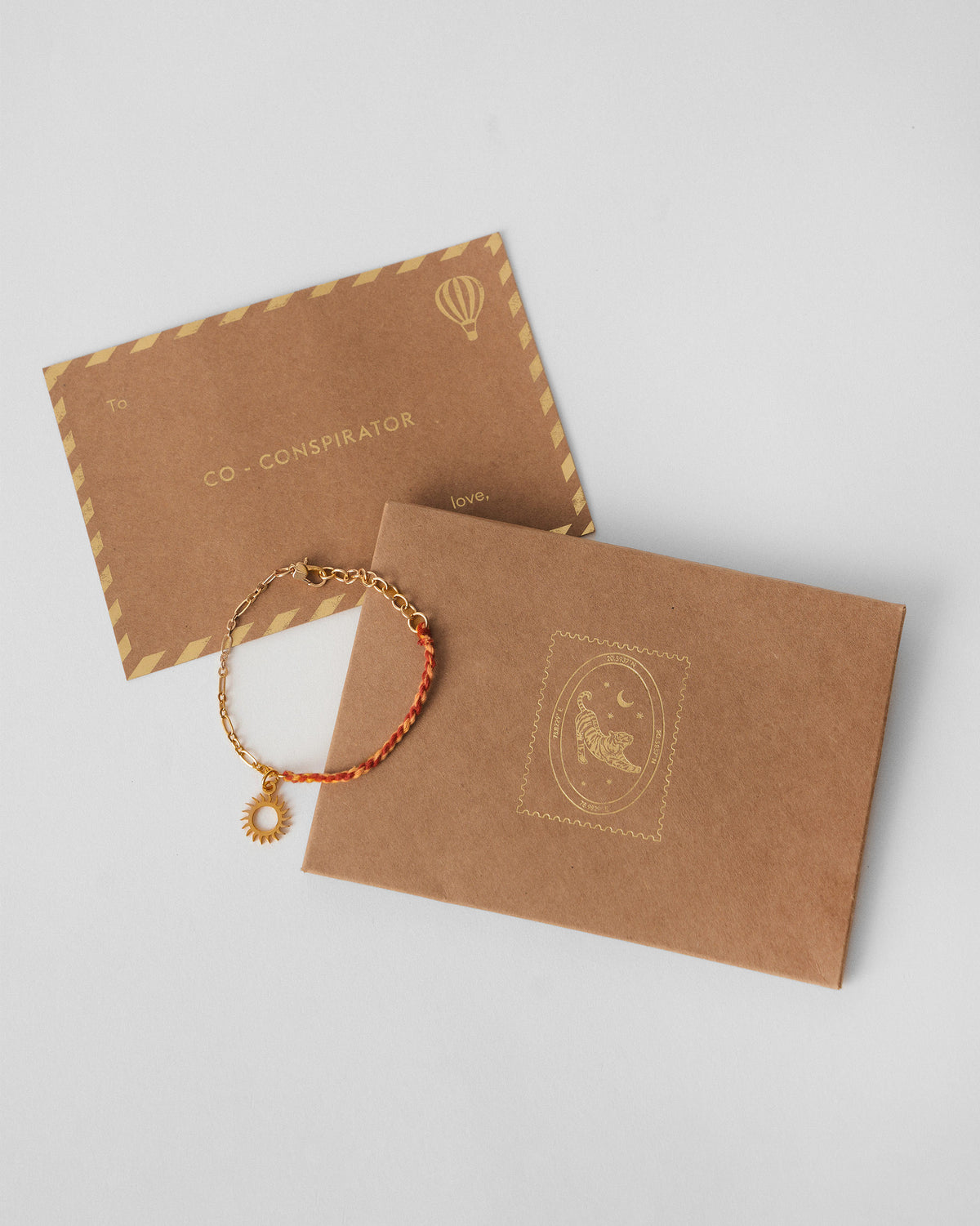 Gold and red Sun Charmlet laid beside a kraft gift envelope and card.