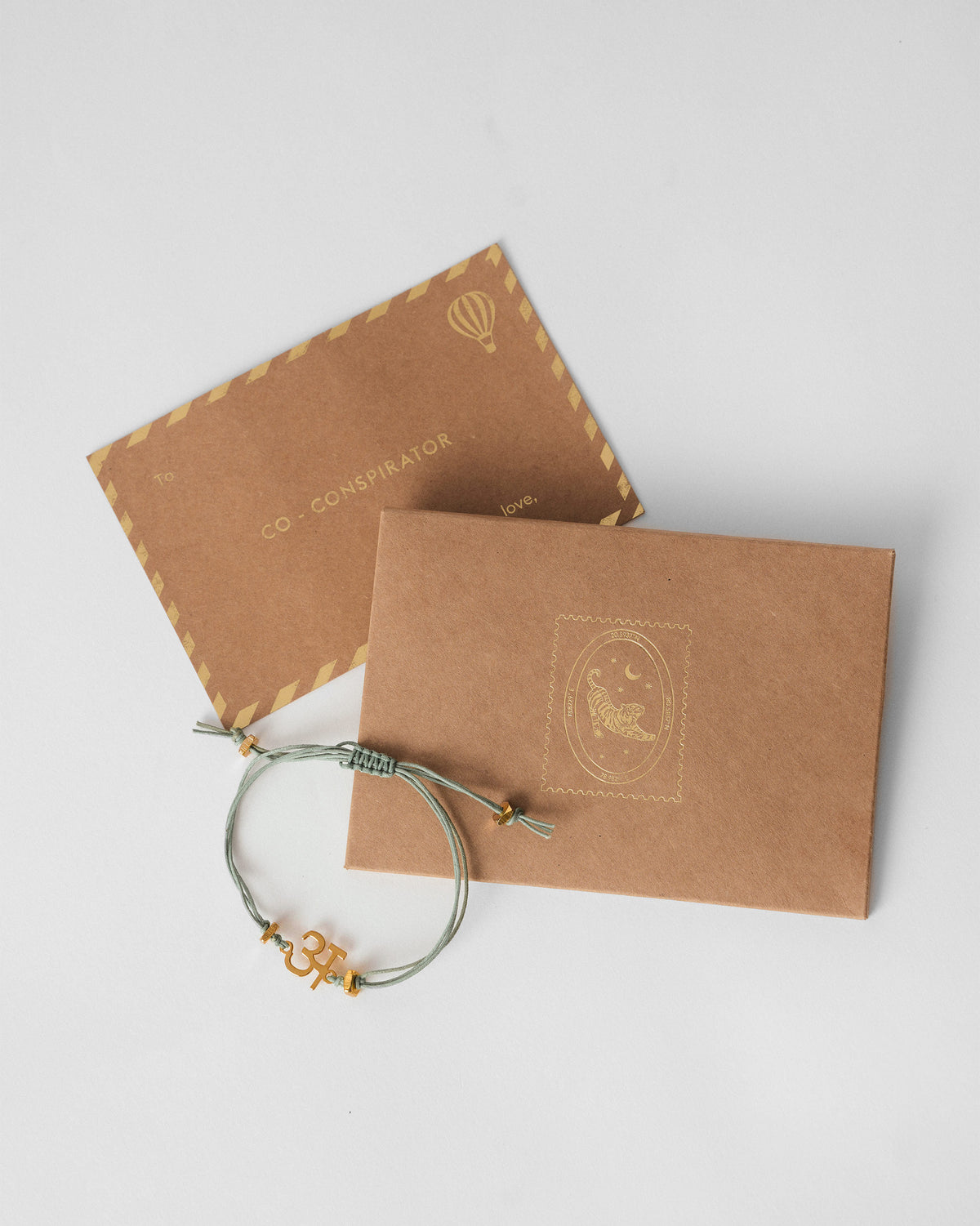Aqua alphabet rakhi with a gold charm beside kraft gift envelopes with gold foil.