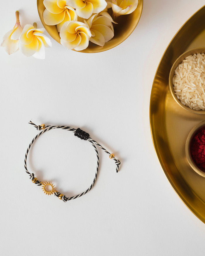 Surya Rakhi in charcoal and white stripes with a gold sun charm, laid flat.