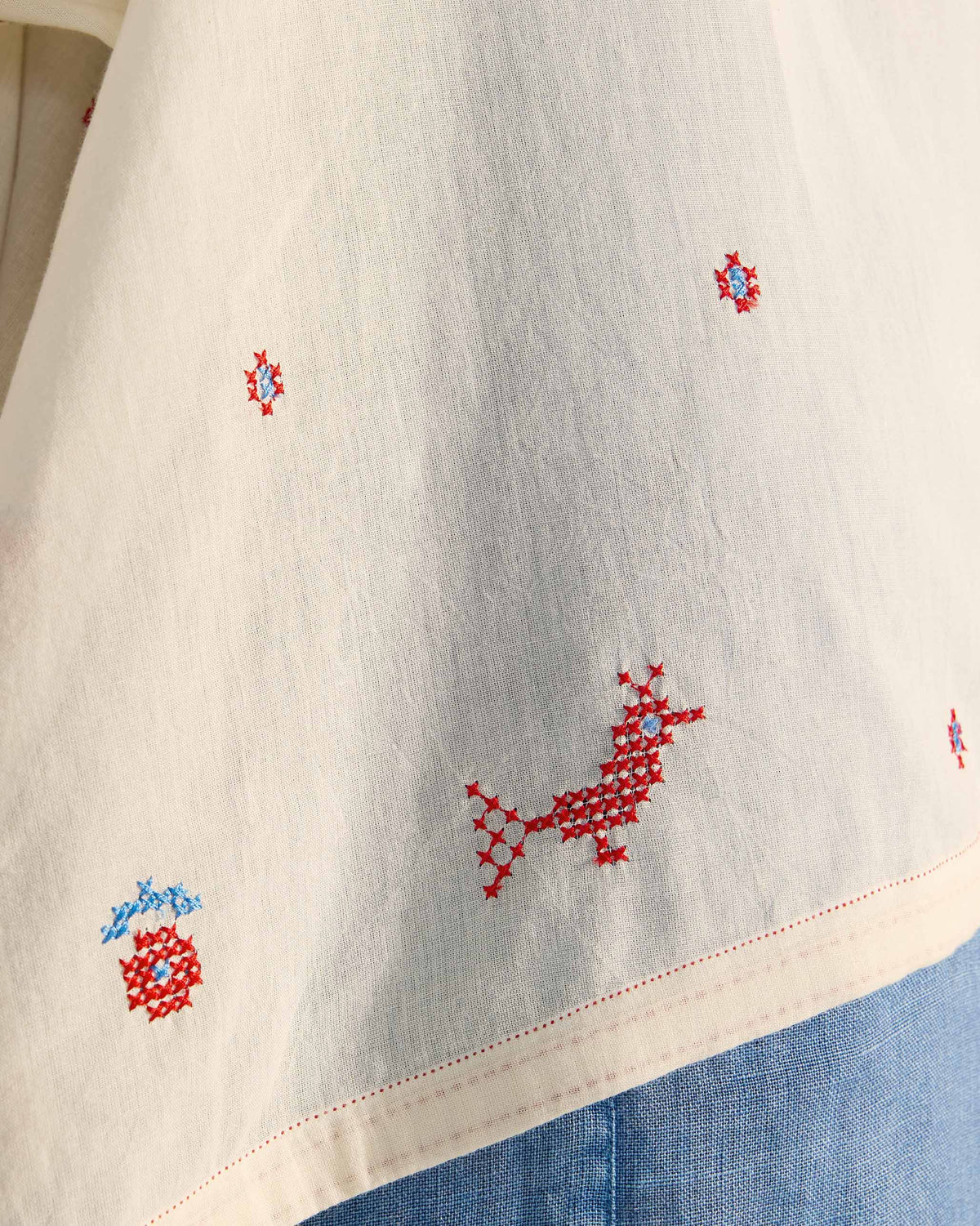 Close-up of red and blue cross-stitch motifs on an ivory Tahi top.