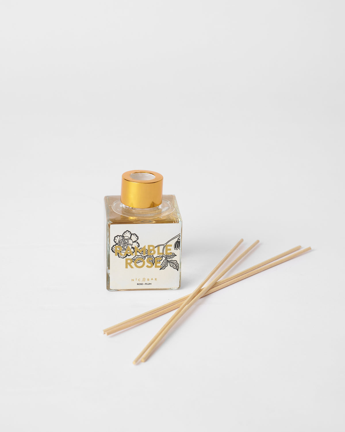 Ramblerose reed diffuser with a gold cap and reeds laid beside it.