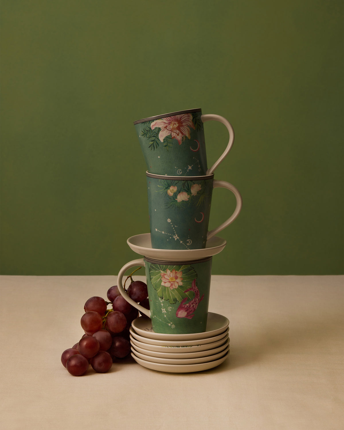 Stack of Nico Pisces mugs on saucers, with grapes beside them.