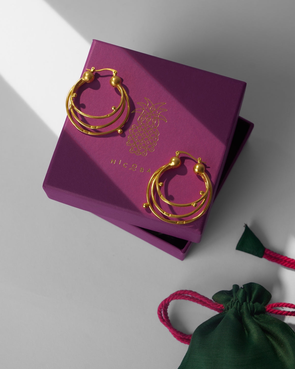 Gold Celestial balis on a purple Nicobar gift box beside a green pouch.