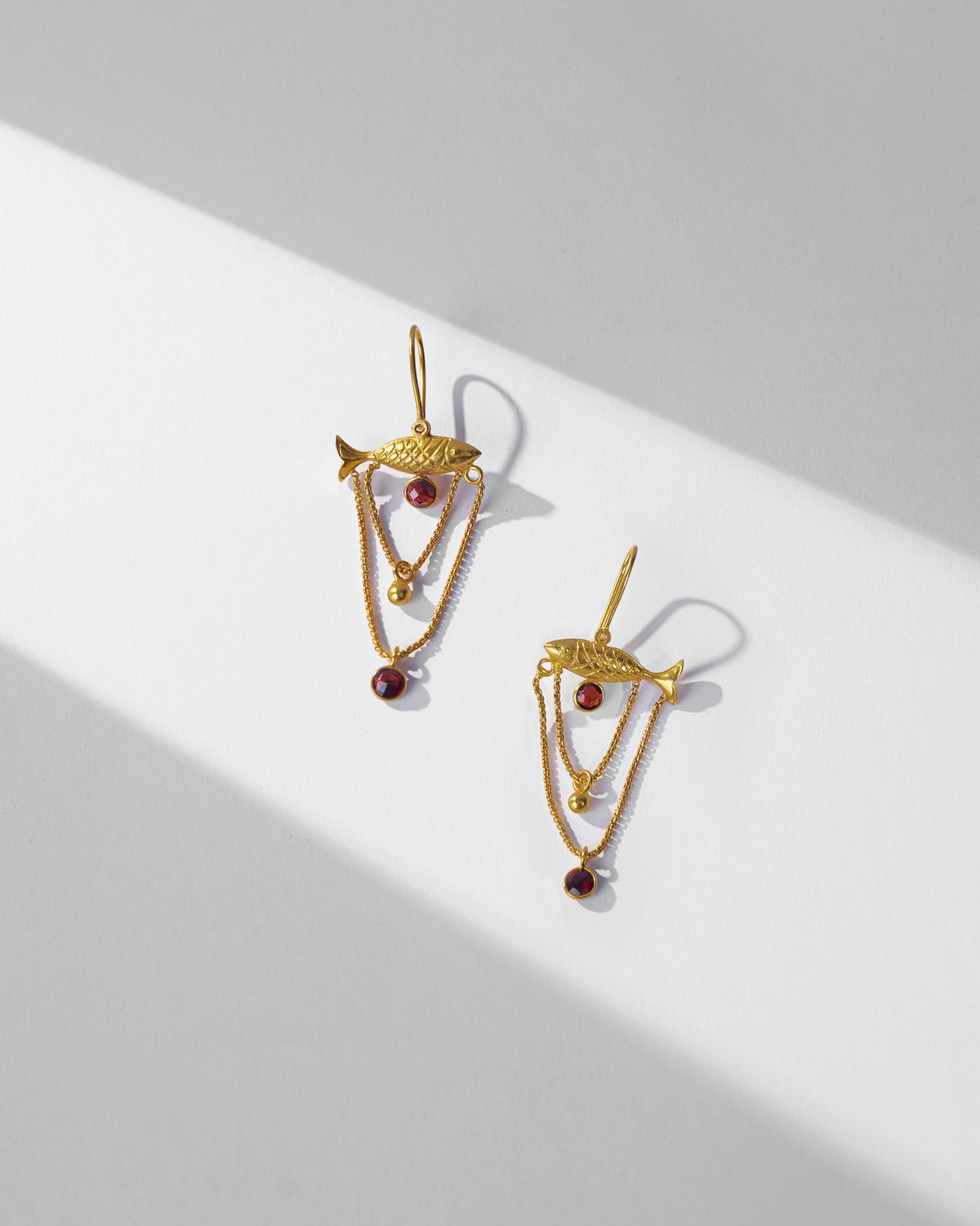 Pair of gold Maase fish earrings with red glass drops laid flat.