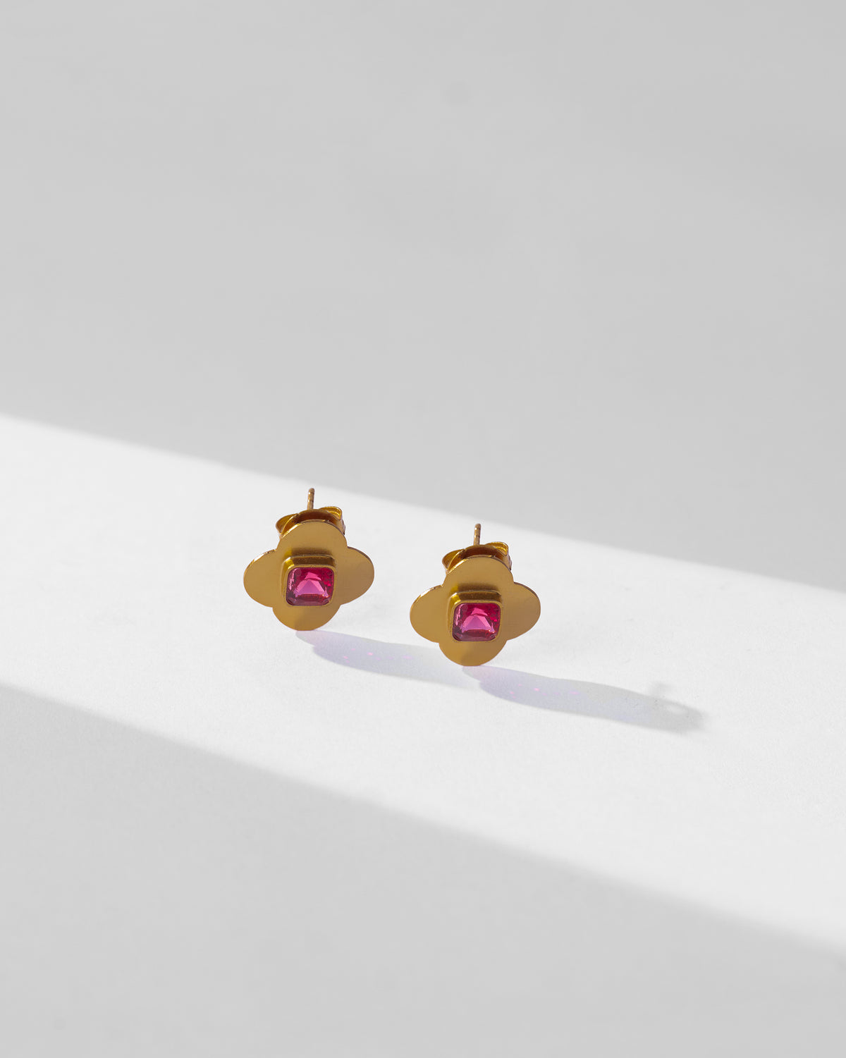 Pair of gold Ahiliya studs with pink glass quatrefoil centres laid flat.