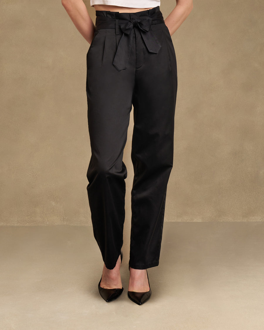 The Gumdrop Pants, front view: black cotton satin narrow-fit trousers with a high waist tied in a bow, worn with black heels.