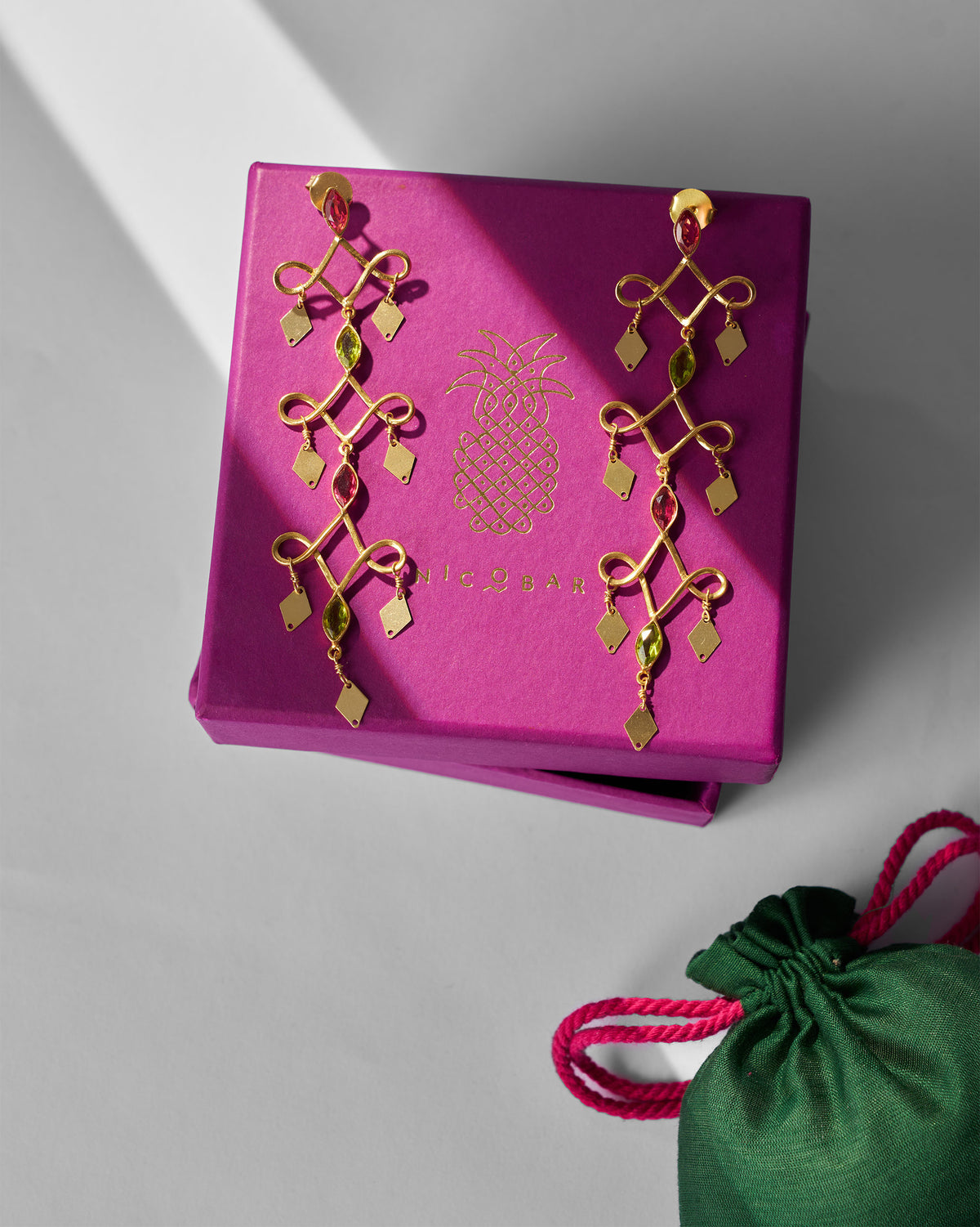 Pair of gold Kolam Danglers on a pink gift box beside a green pouch.