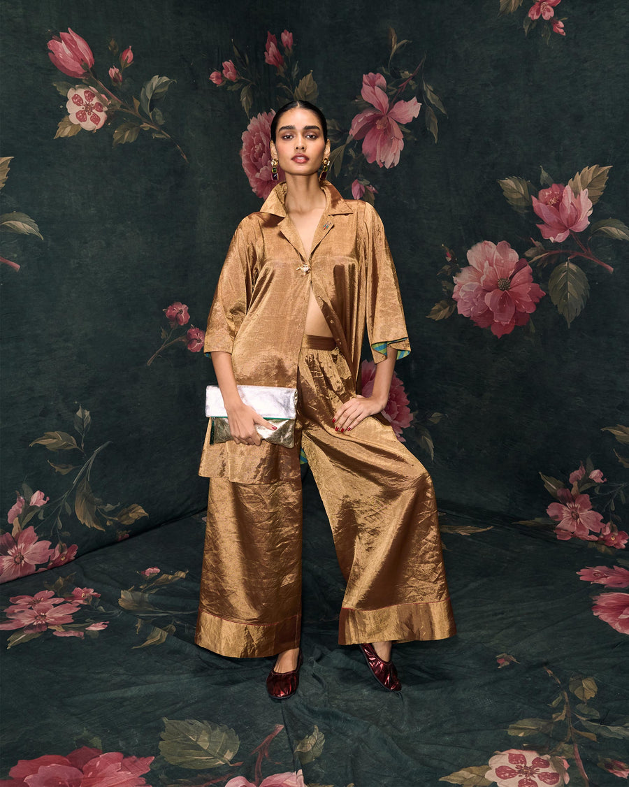 The Golden Hour Shirt worn over gold Charisma Pants with a clutch, against a dark floral backdrop.