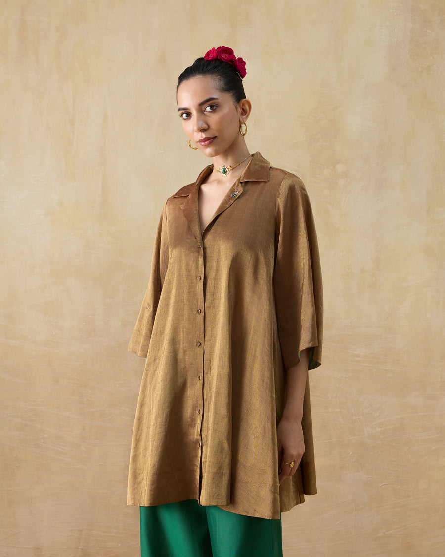 The Golden Hour Shirt, a long gold metallic tissue shirt with a camp collar and flare sleeves, worn over green trousers.