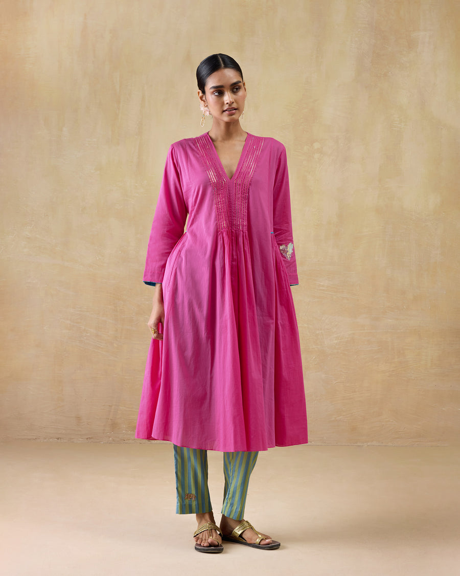 The Pintuck Kurta, a long bright pink cotton kurta with a pintucked V-neck and a gathered waist, worn over striped trousers.