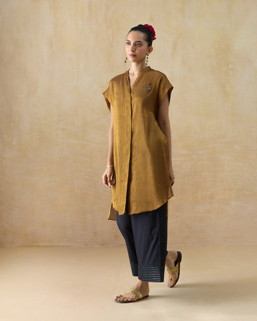 The Tango Kaftan, a gold metallic tissue kaftan with a band collar and cap sleeves, worn over dark trousers.