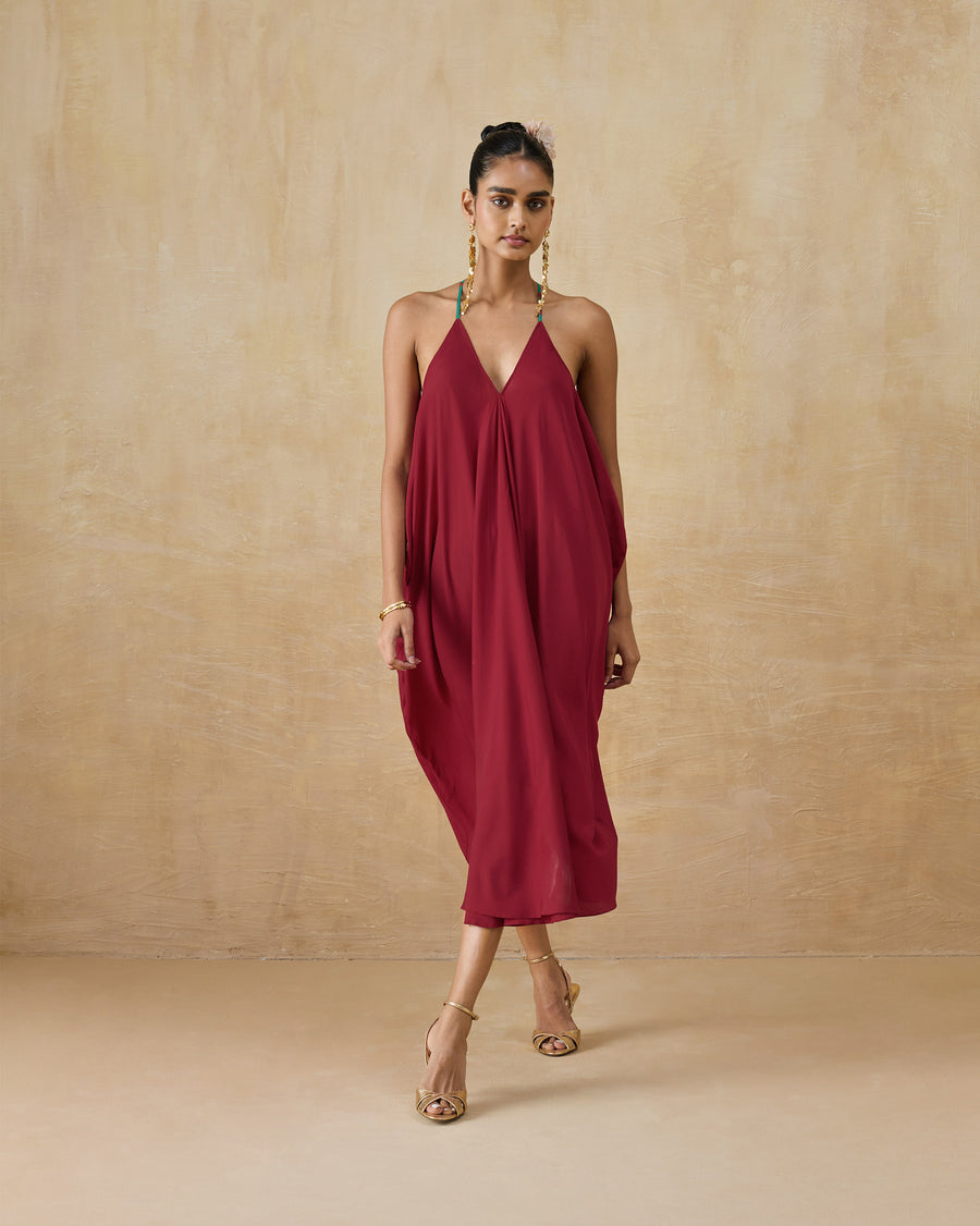 The Savni Dress, a mid-length wine red slip dress with a V-neck and thin straps, worn with gold heels.