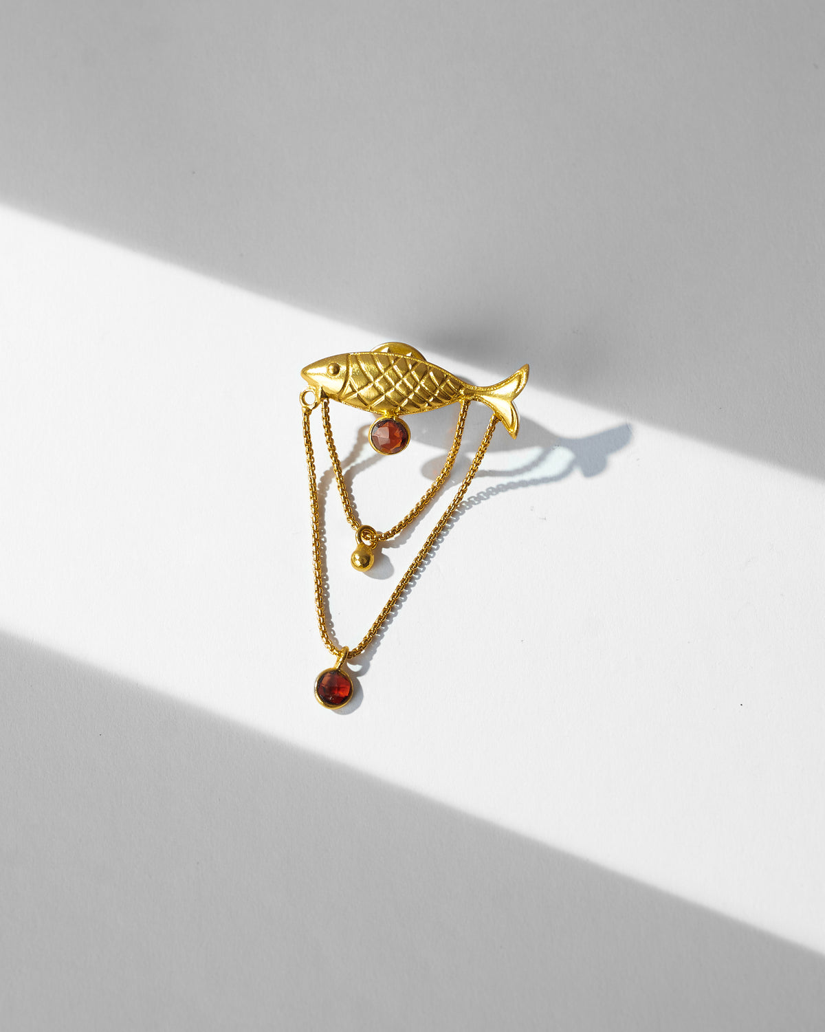 Gold Masse Pin with a fish motif and dangling chains laid flat.