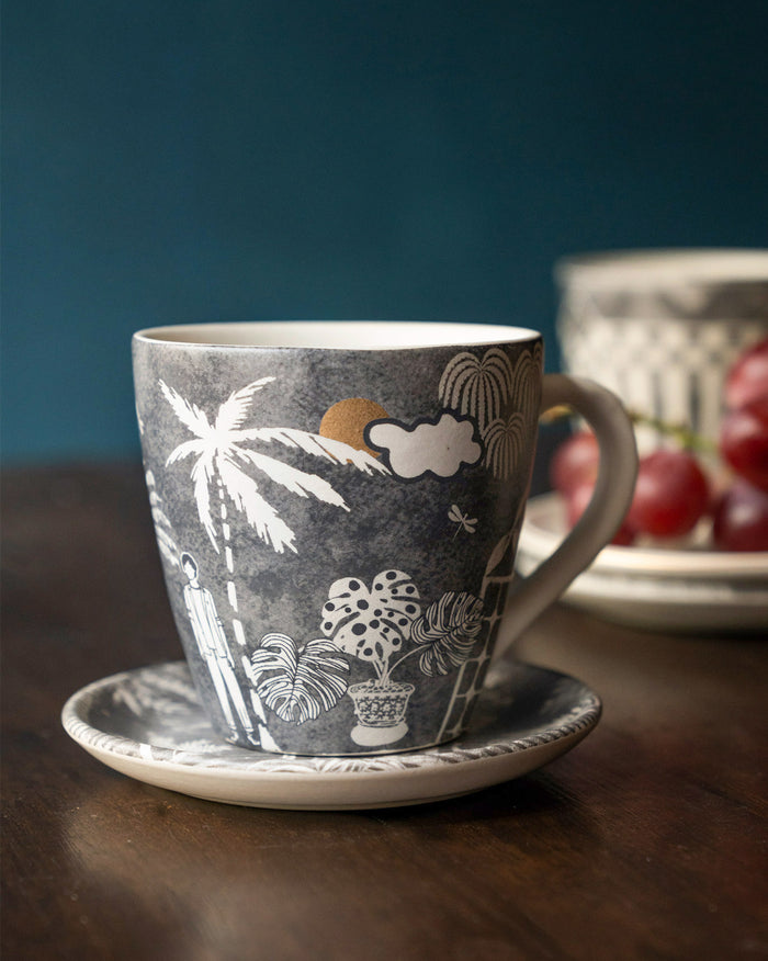 Mandovi Palm Mug in charcoal with white palm and monstera prints, on a saucer.