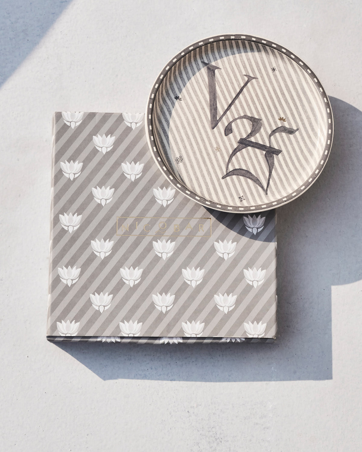 Akshar Coaster - V resting on its grey lotus-print box.