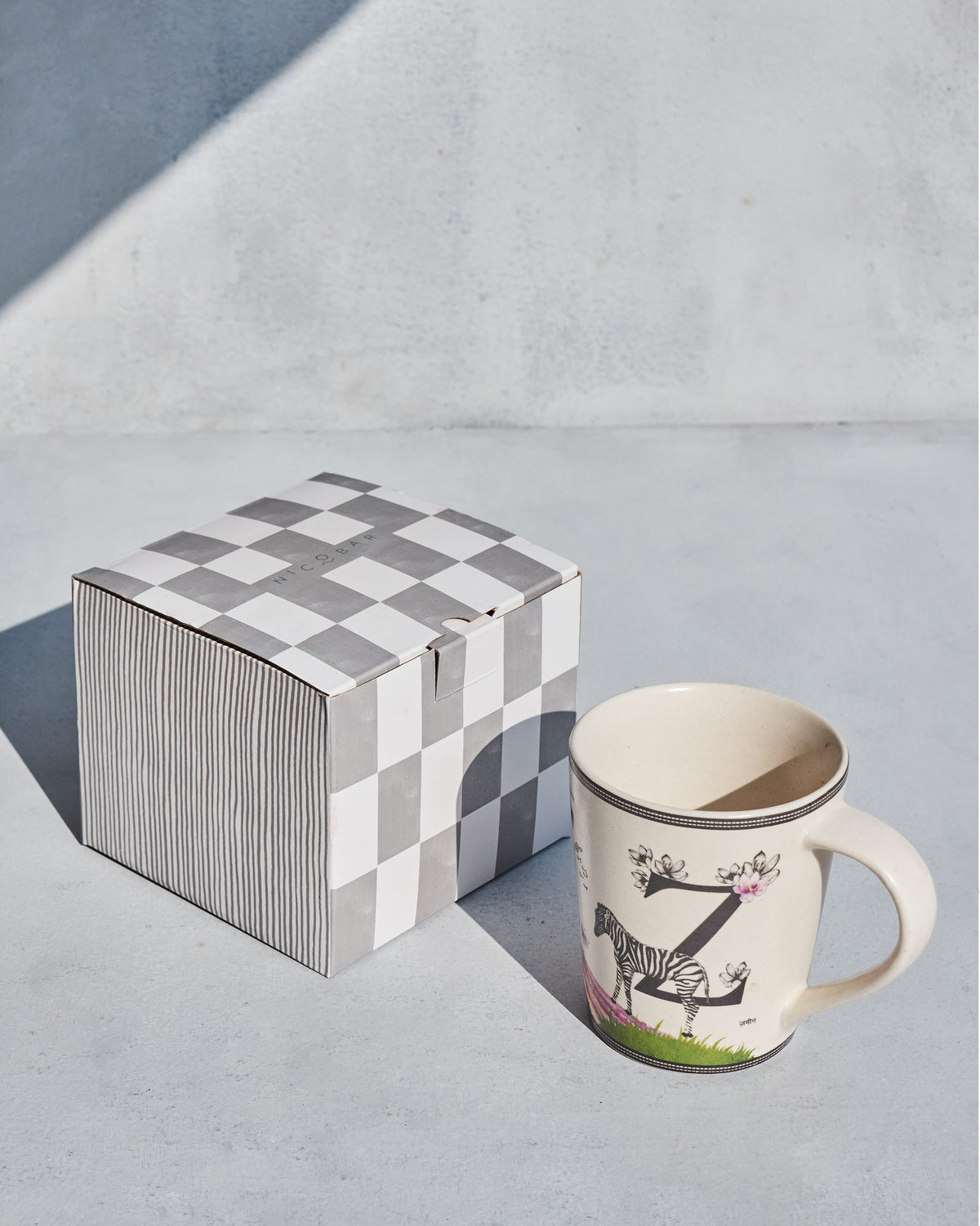 Cream Zafran alphabet mug beside a grey-and-white checked Nicobar gift box.
