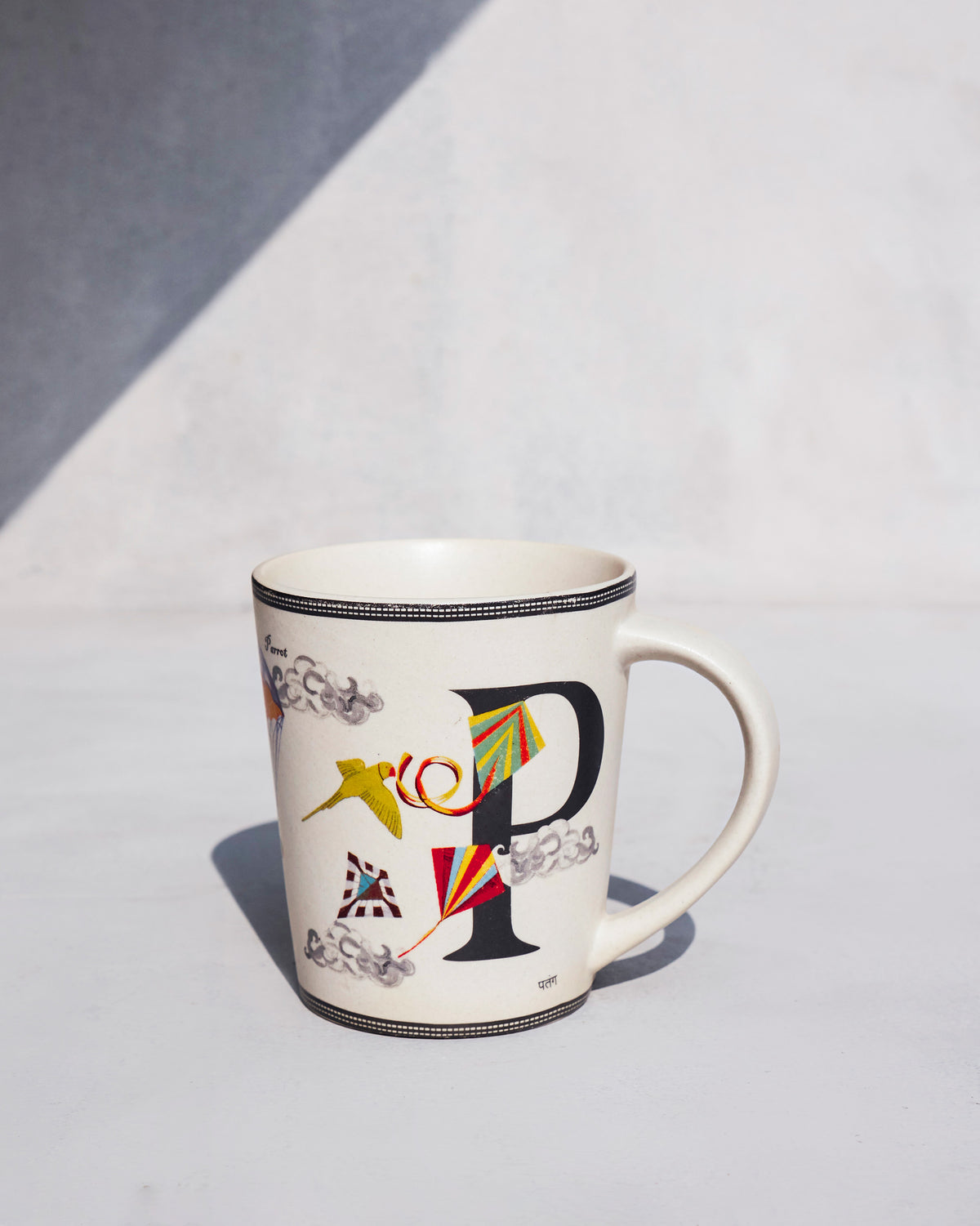 Patang mug with letter P, striped kites, and a green parrot on cream stoneware.