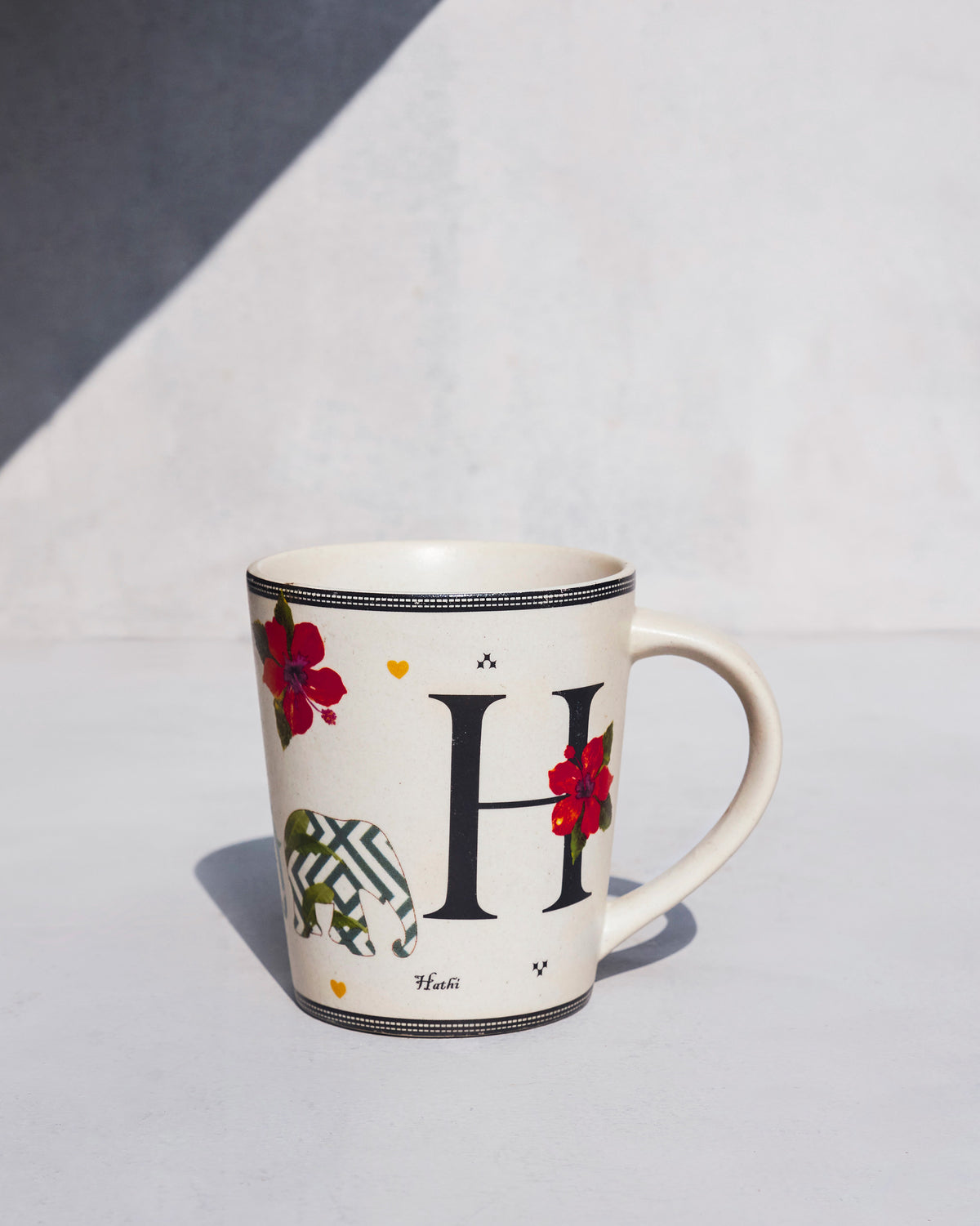 Cream Hans mug with a black letter H, hibiscus flowers, and a patterned elephant.